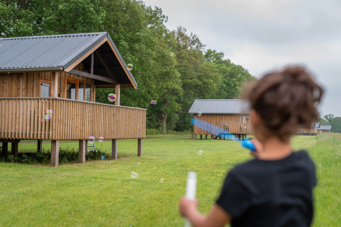 Niño juega con burbujas frente a cabañas de madera en Ackerlodges Ruinerwold, parque de vacaciones en Drenthe.