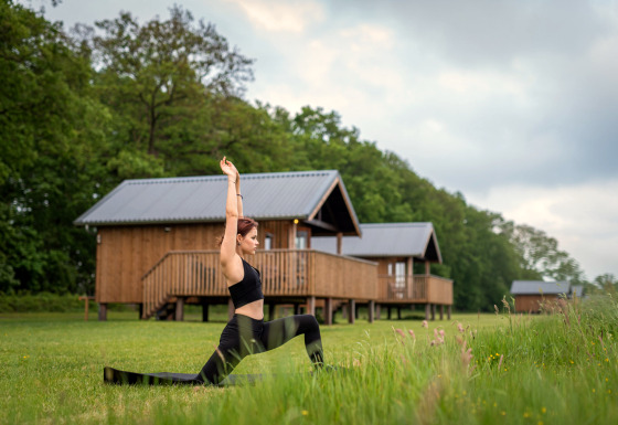 Femme pratiquant le yoga devant Ackerlodge aux Ackerlodges Ruinerwold BV, Pays-Bas, avec des cabanes derrière.