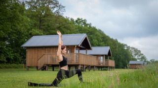 Vrouw doet yoga voor Ackerlodge bij Ackerlodges Ruinerwold BV in Nederland, met houten huisjes op de achtergrond.