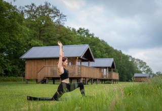 Vrouw doet yoga voor Ackerlodge bij Ackerlodges Ruinerwold BV in Nederland, met houten huisjes op de achtergrond.