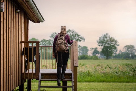 Woman with backpack climbs steps to a wooden cabin at Ackerlodges Ruinerwold BV, Drenthe, Netherlands.