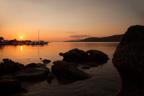 Tramonto sul lago presso un parco vacanze con barche, rocce e sistemazioni glamping sullo sfondo.