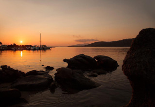 Tramonto sul lago presso un parco vacanze con barche, rocce e sistemazioni glamping sullo sfondo.