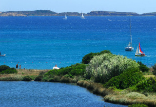View of a glamping holiday park by the sea with green bushes, people on the beach, and sailboats on the water.