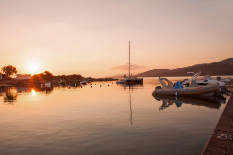 Atardecer en un puerto tranquilo con barcos, autocaravana y zona de glamping en un entorno natural.