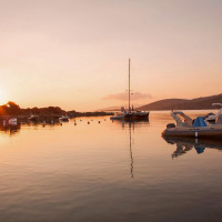 Atardecer en un puerto tranquilo con barcos, autocaravana y zona de glamping en un entorno natural.