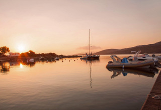 Sunset over a peaceful harbor with boats, camper vans, and glamping area set in a scenic landscape.