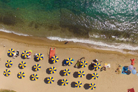 Aerial view of a sandy beach with rows of blue and yellow umbrellas and clear water at a holiday park.