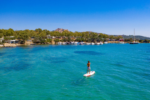 Persona practicando paddle surf en aguas azules frente a un parque de glamping con barcos y árboles.