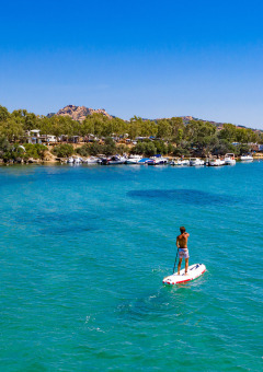 Persona practicando paddle surf en aguas azules frente a un parque de glamping con barcos y árboles.