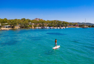 Stand up paddle boarder on blue water near a glamping holiday park with boats, trees and clear sky.