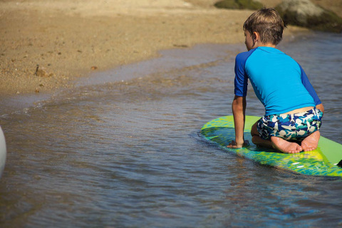 Dreng i blå rashguard og badebukser knæler på et surfbræt ved lavvandet strand ved feriepark.