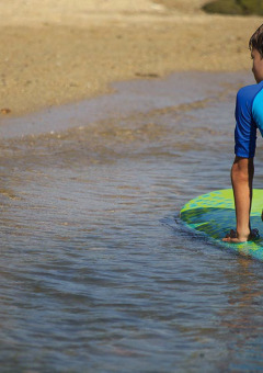 Niño con camiseta azul arrodillado en una tabla de surf junto al agua en un parque de vacaciones glamping.