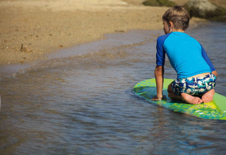 Niño con camiseta azul arrodillado en una tabla de surf junto al agua en un parque de vacaciones glamping.