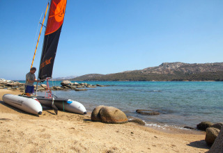 Uomo prepara un catamarano sulla spiaggia sabbiosa di un villaggio vacanze, con mare azzurro e colline sullo sfondo.