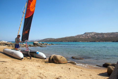 Uomo prepara un catamarano sulla spiaggia sabbiosa di un villaggio vacanze, con mare azzurro e colline sullo sfondo.