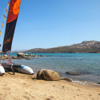 Hombre preparando un catamarán en la playa arenosa de un parque vacacional, con mar azul y colinas detrás.