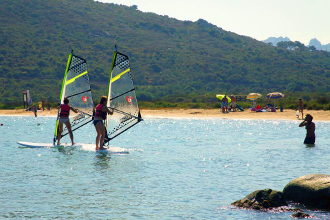 Two people are windsurfing near a beach at a holiday park with glamping tents, sun umbrellas, and swimmers.