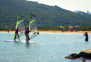 Two people are windsurfing near a beach at a holiday park with glamping tents, sun umbrellas, and swimmers.