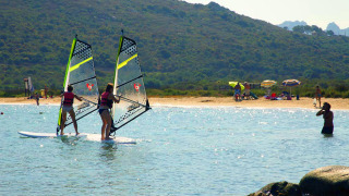 Dos personas practican windsurf cerca de una playa con sombrillas y tiendas de glamping en un parque vacacional.