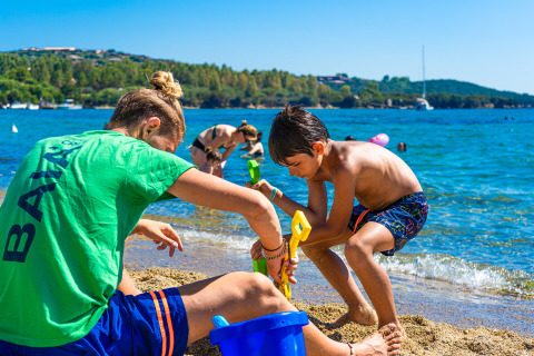 Dos niños juegan en la arena junto al agua en un parque vacacional con glamping, rodeados de naturaleza.