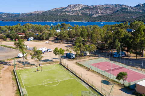 Vista aerea di un parco vacanze glamping con campi da tennis e calcio, alberi, lago e montagne.