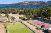 Vista aerea di un parco vacanze glamping con campi da tennis e calcio, alberi, lago e montagne.