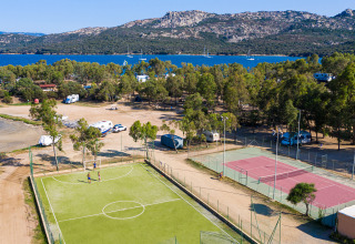 Aerial view of a holiday park with glamping, tennis and football courts, trees, lake, and mountains.