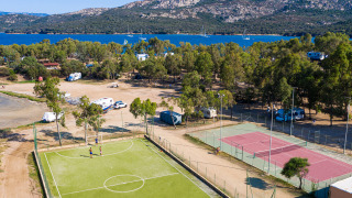 Vista aérea de parque vacacional con glamping, canchas de tenis y fútbol, árboles, lago y montañas.