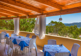 Udendørs restaurantterrasse med blådugede borde, hvide stole og naturskøn udsigt i feriepark.