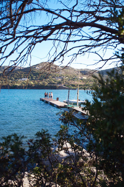 Vista a través de árboles de un lago, muelle con personas y colinas al fondo en un parque de glamping.