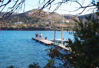 Vista tra gli alberi di un lago, molo con persone e colline sullo sfondo in un parco vacanze glamping.