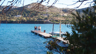 Vista a través de árboles de un lago, muelle con personas y colinas al fondo en un parque de glamping.