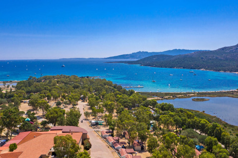 Vista aérea de un parque vacacional con árboles, laguna azul y barcos en la costa junto al mar en Italia.