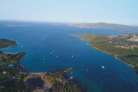 Vista aérea de un parque vacacional con glamping junto al mar, rodeado de bosques, playas y veleros.