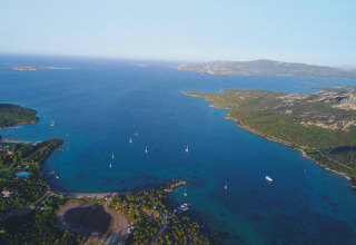 Vista aérea de un parque vacacional con glamping junto al mar, rodeado de bosques, playas y veleros.