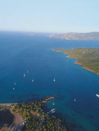 Vista aérea de un parque vacacional con glamping junto al mar, rodeado de bosques, playas y veleros.