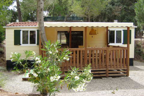 SunLodge Mobile Home Redwood en Camping Village Capo d'Orso, Italia, rodeado de árboles y flores blancas.