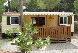 SunLodge Mobile Home Redwood en Camping Village Capo d'Orso, Italia, rodeado de árboles y flores blancas.