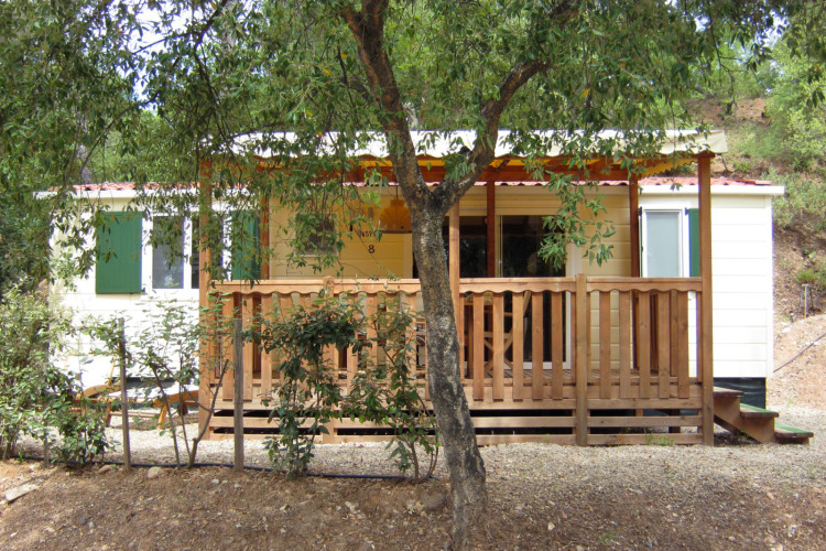SunLodge Mobile Home Aspen en Camping Village Capo d'Orso, Italia, con terraza de madera y árboles delante.