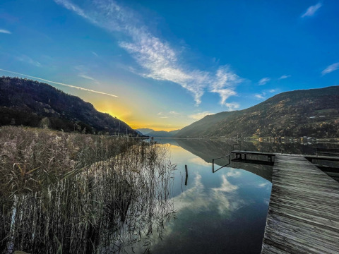 Sunset over a calm lake with mountains, reeds and a wooden pier, taken at a glamping holiday park.
