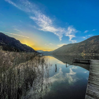 Atardecer sobre un lago tranquilo con montañas y muelle, visto desde un parque de glamping vacacional.