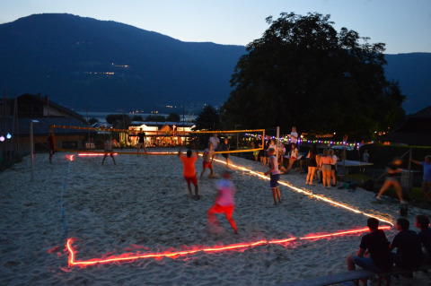 Partido de voleibol en la arena al atardecer en un parque vacacional con glamping, luces y montañas.