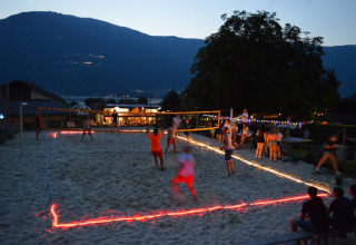 Aftenbillede af strandvolleyball på en ferielejrplads med glamping, omgivet af bjerge og lysguirlander.