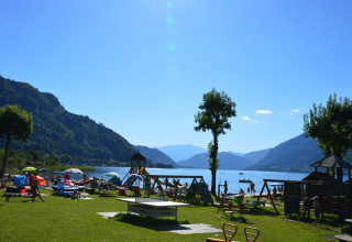 Sommerdag ved søbred med folk, legeplads, paraplyer og bjerge i baggrunden i en feriepark med glamping.