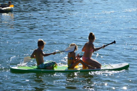 Tres niños reman en una tabla sobre un lago soleado durante unas vacaciones en un parque glamping.
