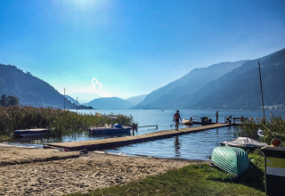 Sunny day by a calm lake at a holiday park with dock, boats, and people, surrounded by mountains.