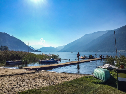 Zonnige dag aan een rustiek meer in een vakantiedomein met steiger, boten en bergen op de achtergrond.