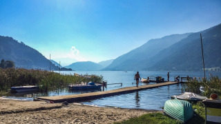 Día soleado junto a un lago en un parque de vacaciones, con muelle, barcos, gente y montañas alrededor.