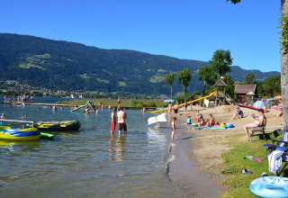 Families relaxing and swimming at a lakeside holiday park with mountains, offering glamping accommodations.